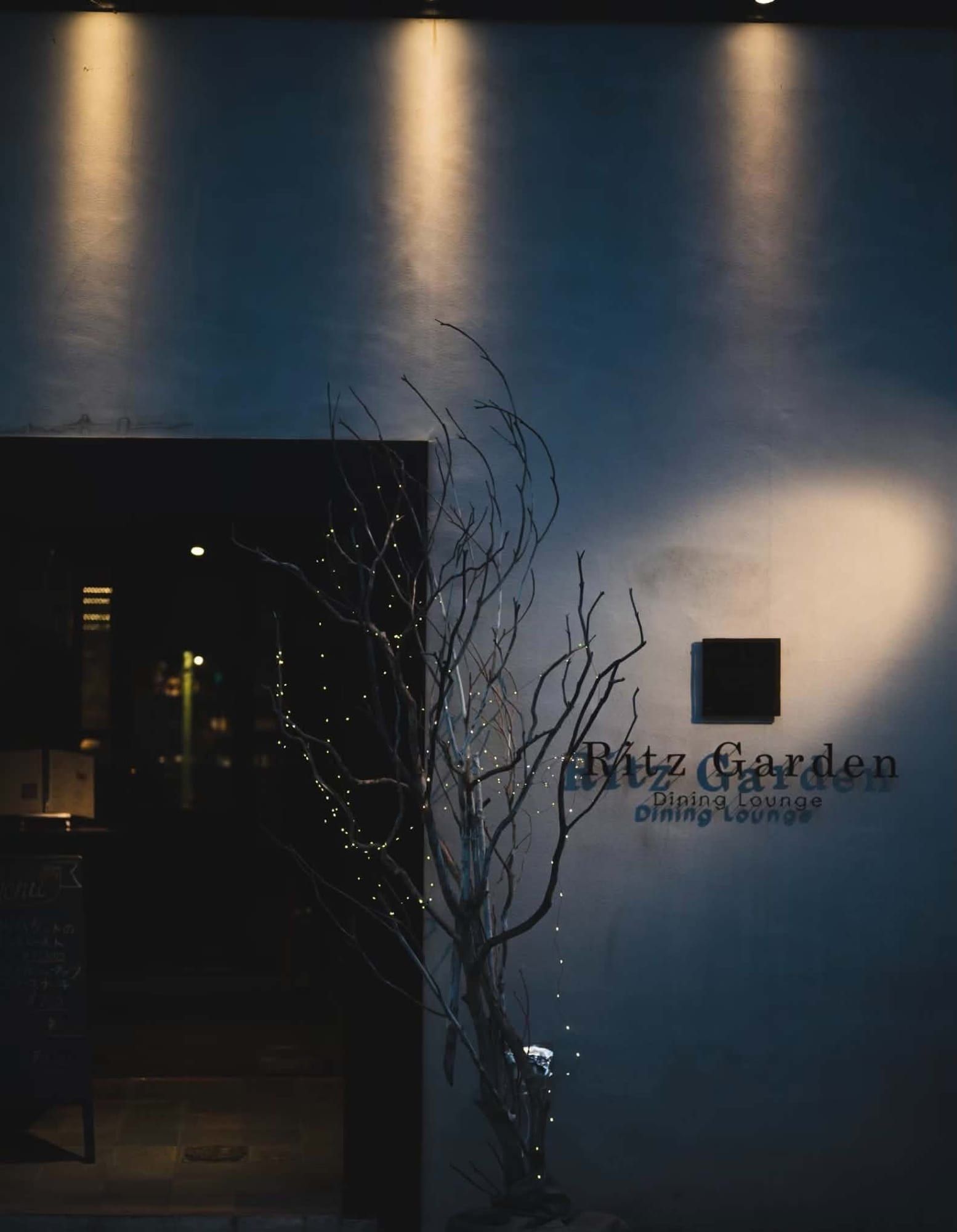 夜裡亮起的「Ritz Garden Dining Lounge」招牌，旁邊是纏著燈飾的枝條